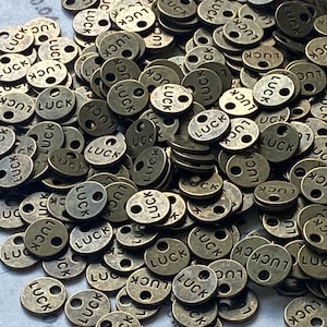 May include: A collection of small, round, bronze-colored metal charms. Each charm has the word "LUCK" stamped on it, along with two small holes. The charms are scattered, creating a textured surface.