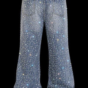 May include: Light blue denim jeans heavily embellished with sparkling rhinestones. The jeans have a flared leg design and feature classic five-pocket styling. The rhinestones are densely applied, creating a shimmering effect across the entire garment.