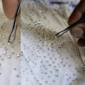 May include: Close-up of a white fabric densely covered with small, sparkling rhinestones. A pair of tweezers is used to handle the rhinestones, suggesting a craft or embellishment project. The rhinestones are evenly spaced across the fabric.