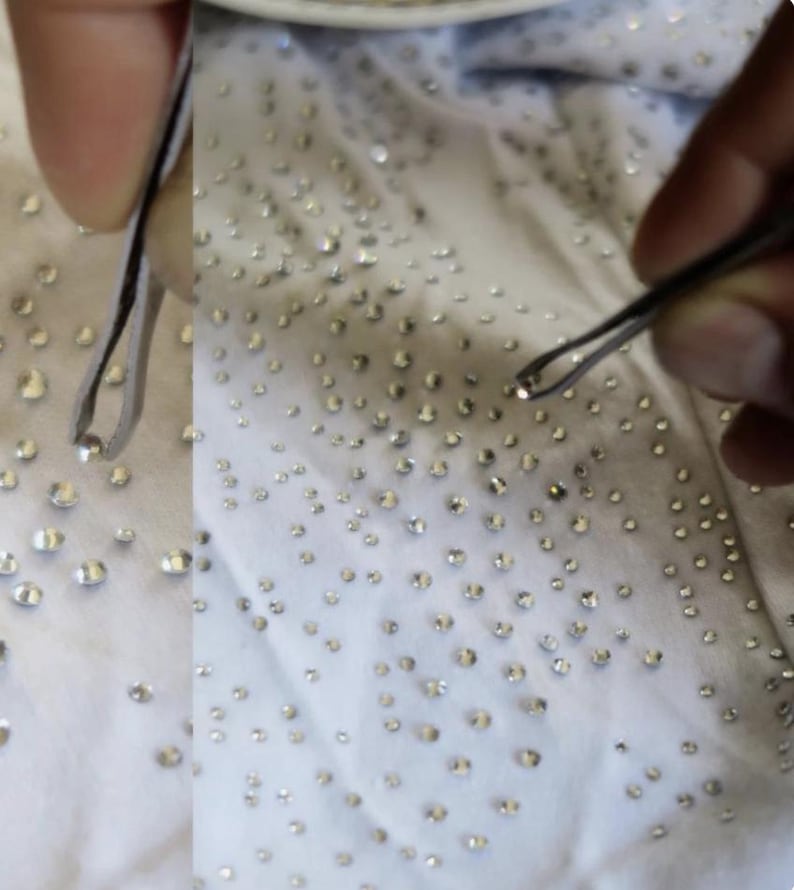 May include: Close-up of a white fabric densely covered with small, sparkling rhinestones. A pair of tweezers is used to handle the rhinestones, suggesting a craft or embellishment project. The rhinestones are evenly spaced across the fabric.