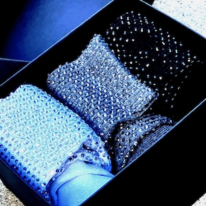 May include: A black box containing several folded, shimmering scarves in shades of blue. The scarves have a textured, sequined appearance, with varying patterns and densities of embellishments. The box is a deep navy blue.