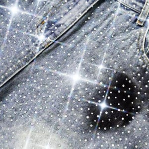 May include: Close-up of denim jeans heavily embellished with sparkling rhinestones. The light blue jeans have a classic design with pockets and a waistband. The rhinestones are evenly distributed across the fabric, creating a dazzling effect.