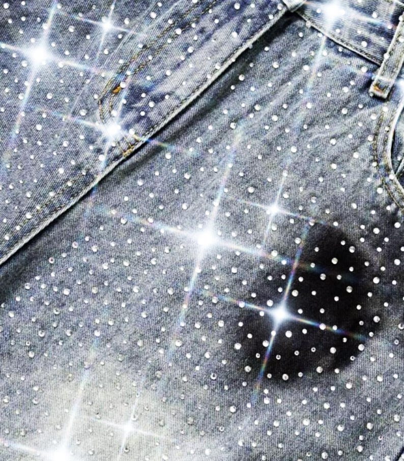 May include: Close-up of denim jeans heavily embellished with sparkling rhinestones. The light blue jeans have a classic design with pockets and a waistband. The rhinestones are evenly distributed across the fabric, creating a dazzling effect.