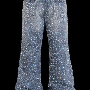 May include: Light blue denim jeans heavily embellished with sparkling rhinestones. The jeans have a classic five-pocket design and a flared leg. The rhinestones are densely packed, creating a shimmering effect.