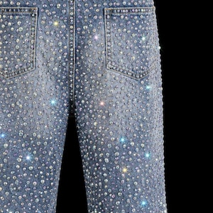 May include: Light blue denim jeans heavily embellished with sparkling rhinestones. The jeans feature classic five-pocket styling and a straight leg cut. The back pockets are visible, and the overall effect is glamorous and eye-catching.
