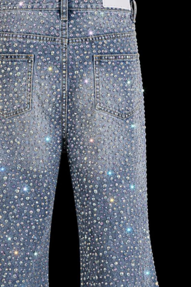 May include: Light blue denim jeans heavily embellished with sparkling rhinestones. The jeans feature classic five-pocket styling and a straight leg cut. The back pockets are visible, and the overall effect is glamorous and eye-catching.