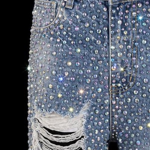May include: Light blue denim jeans heavily embellished with iridescent rhinestones. The jeans feature a distressed, ripped knee detail. The overall effect is a sparkling, glamorous look.