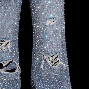 May include: Light blue denim jeans heavily embellished with sparkling rhinestones. The jeans feature distressed details with ripped sections, creating a glamorous and edgy aesthetic. The rhinestones are of varying sizes.