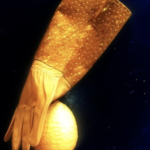 May include: A tan leather glove with a dotted brown cuff rests next to a textured, yellow lemon. The glove's design features a long cuff and a smooth, fitted palm. The background is a dark blue.