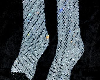 May include: A pair of sheer, light-coloured socks densely covered in sparkling rhinestones. The toe and heel sections are black, creating a contrast with the shimmering body of the socks. The socks are displayed against a dark background.