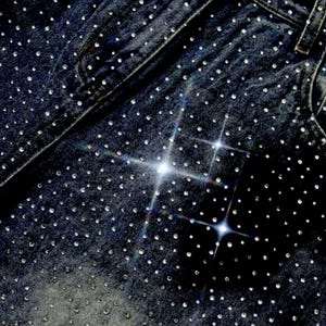 May include: Close-up of dark wash denim jeans heavily embellished with small, sparkling rhinestones. The rhinestones are evenly distributed across the fabric, creating a shimmering effect. The jeans feature classic pocket and seam details.