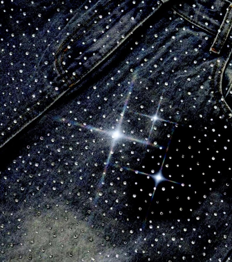 May include: Close-up of dark wash denim jeans heavily embellished with small, sparkling rhinestones. The rhinestones are evenly distributed across the fabric, creating a shimmering effect. The jeans feature classic pocket and seam details.