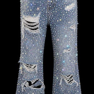 May include: Light blue denim jeans with a flared leg design, heavily embellished with iridescent rhinestones. The jeans feature distressed details with ripped sections throughout the legs, creating a sparkling, edgy look.