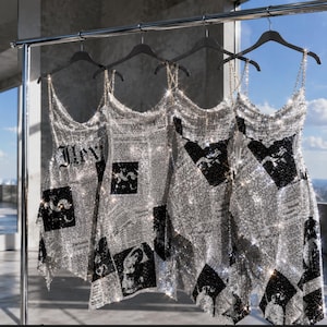 May include: Four sleeveless dresses hanging on a metal rack, each crafted from shimmering silver metallic mesh. The fabric features a black and white newspaper-style print with various graphic illustrations and text blocks, suspended by thin chain straps.