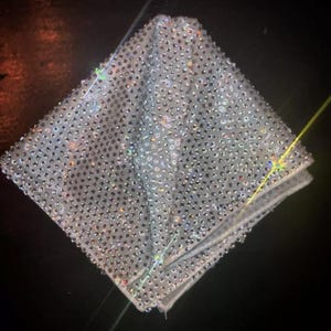 May include: A square, folded, silver-colored fabric, densely covered with small, sparkling rhinestones. The fabric is folded into a diamond shape, with the edges visible. The rhinestones catch the light, creating a shimmering effect.