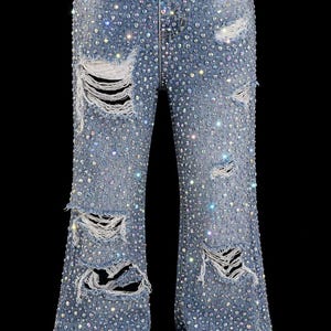 May include: Light blue denim jeans with distressed details and embellished with numerous sparkling rhinestones. The jeans have a flared leg design and are set against a black background.
