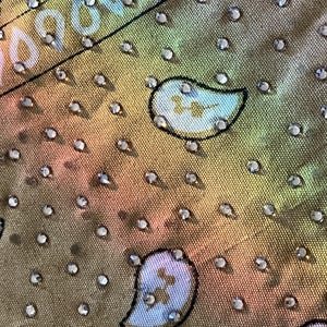 May include: Close-up of a fabric with a paisley pattern in shades of gold, pink, and green. The fabric is embellished with numerous small, sparkling rhinestones, adding a touch of glamour and texture to the design.
