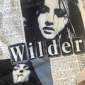 May include: A white fabric garment featuring a black and white graphic print of two faces and newspaper-style text columns. The word Wilder is printed in large, bold, white capital letters across a black rectangular background in the centre.