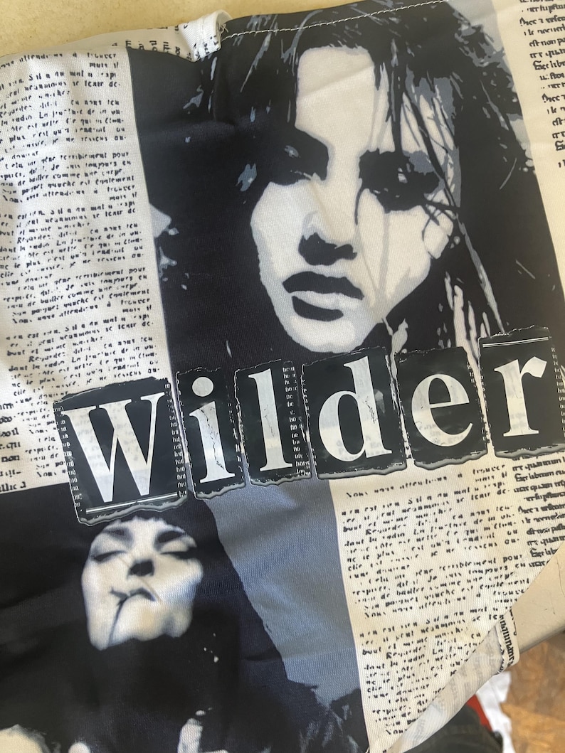 May include: A white fabric garment featuring a black and white graphic print of two faces and newspaper-style text columns. The word Wilder is printed in large, bold, white capital letters across a black rectangular background in the centre.