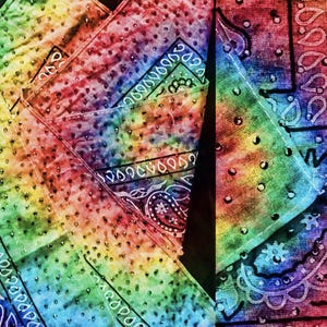 May include: Close-up of several rainbow-colored bandanas. Each bandana features a tie-dye pattern with a variety of colors, including red, orange, yellow, green, blue, and purple. The bandanas have a paisley design and a decorative border.