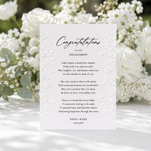 May include: A white rectangular engagement card featuring embossed floral borders and black printed text that reads Congratulations on your ENGAGEMENT, followed by a poem and the names LEAH + KANE with the date 24.05.2029 at the bottom.