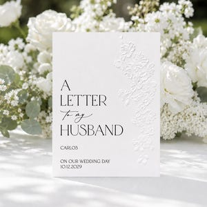 May include: A white rectangular card featuring embossed floral details on the right side and black text that reads A LETTER To my HUSBAND, CARLOS, ON OUR WEDDING DAY, and 10.12.2029. The card has a clean, minimalist design with elegant typography.