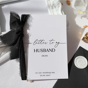 May include: A white rectangular card featuring black text that reads A letter to my HUSBAND DEAN on our wedding day 29.05.2027. The card is decorated with a black frayed-edge ribbon tied along the left side, providing a textured contrast to the paper.