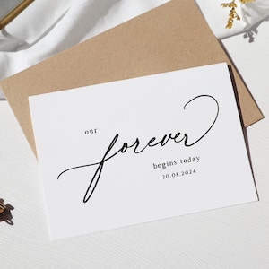 May include: A white card with black calligraphy that reads "our forever begins today 20.08.2024".