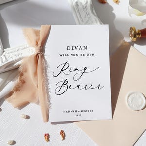 May include: A white card with black text that reads "Devan, will you be our Ring Bearer? Hannah & George 2027". The card is tied with a peach-coloured ribbon.