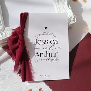 May include: A white card with black text that reads "Congratulations Jessica and Arthur on your wedding day 03.09.2024". A small black heart is above the text. The card is tied with a burgundy ribbon.