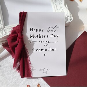 May include: A white card with a red ribbon tied around it. The card says "Happy 1st Mother's Day to my Godmother" with a small heart at the bottom. The card also says "with love from Avery".