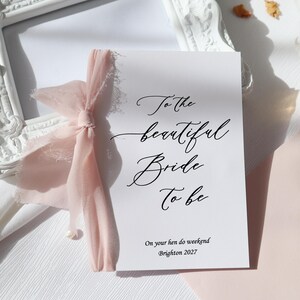 May include: A white card with black calligraphy that reads "To the beautiful bride to be" and "On your hen do weekend Brighton 2027". The card is tied with a pink ribbon.
