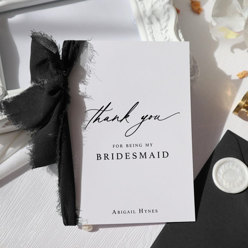 May include: A white card with black text that says "Thank you for being my bridesmaid" and "Abigail Hynes". The card is tied with a black ribbon.