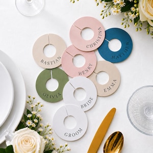 Op de afbeelding: Een verzameling cirkelvormige, platte, ringvormige markers in verschillende kleuren, waaronder wit, beige, roze, olijfgroen en donkerblauw. Elke marker is voorzien van een gleuf en is gepersonaliseerd met zwarte tekst, waaronder namen zoals Bastian, Christina, Matthew, Henry, Emma, Olivia, Bride en Groom.