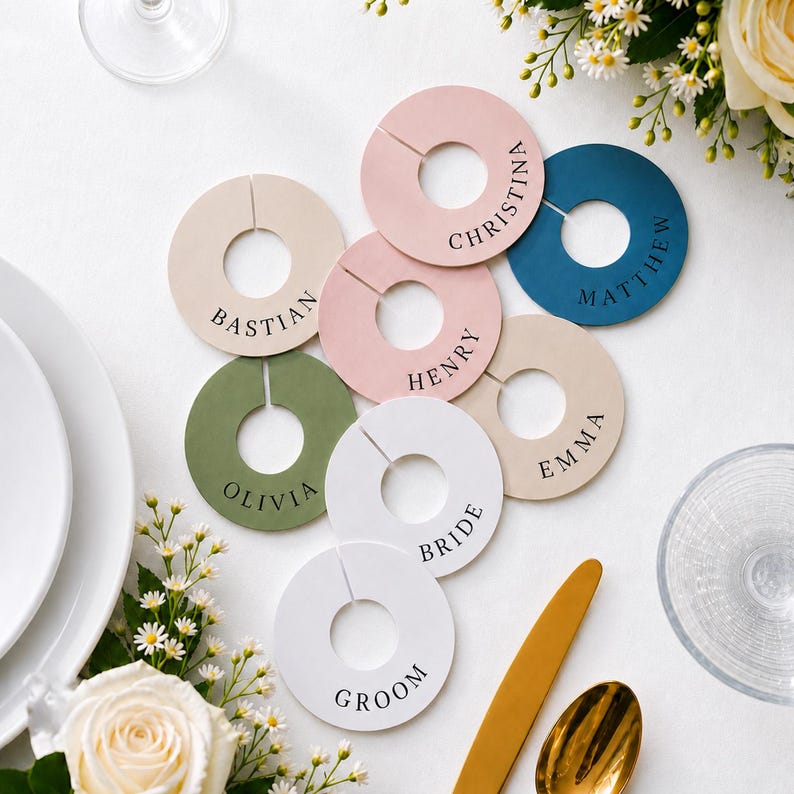 Op de afbeelding: Een verzameling cirkelvormige, platte, ringvormige markers in verschillende kleuren, waaronder wit, beige, roze, olijfgroen en donkerblauw. Elke marker is voorzien van een gleuf en is gepersonaliseerd met zwarte tekst, waaronder namen zoals Bastian, Christina, Matthew, Henry, Emma, Olivia, Bride en Groom.