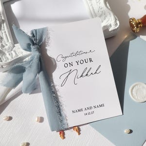 May include: A white rectangular card featuring the black printed text Congratulations ON YOUR Nikkah, NAME AND NAME, and 14.12.27. The card is adorned with a dusty blue frayed silk ribbon tied in a bow on the left side, accompanied by a matching envelope.