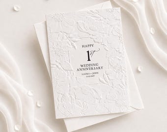 May include: A white greeting card with an embossed floral pattern and black text that reads HAPPY 1ST WEDDING ANNIVERSARY LAURA + JOHN 15.03.2029. The card is placed on top of a matching white envelope, creating a clean and textured appearance.