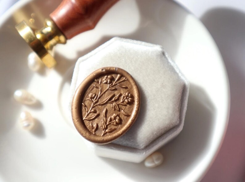 May include: A white velvet ring box with a gold wax seal featuring a floral design. The seal is on top of the box.