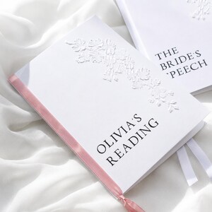 May include: A white notebook with a pink ribbon spine and a white embossed floral design. The text on the cover reads OLIVIA'S READING in black font. A second white notebook is partially visible with the text THE BRIDE'S SPEECH printed in black.