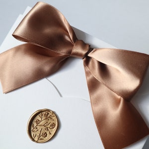 May include: A white envelope with a brown satin ribbon tied in a bow. A gold wax seal with a floral design is on the front of the envelope.
