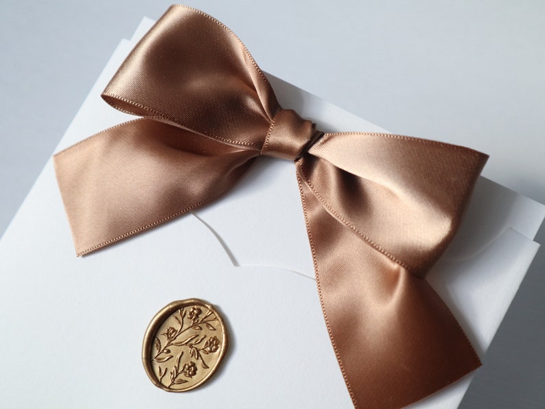 May include: A white envelope with a brown satin ribbon tied in a bow. A gold wax seal with a floral design is on the front of the envelope.