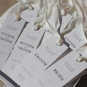 May include: White wedding place cards with black text. The cards are tied with white ribbon and have the text "this seat is kindly reserved for the" followed by the title of the person the seat is reserved for, such as "Father of the Groom" or "Bride".