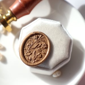 May include: A gold wax seal with a floral design on a white velvet ring box.