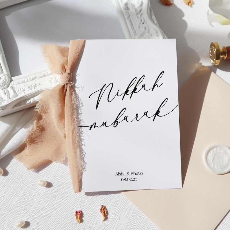 May include: A white rectangular card featuring the black script text Nikkah Mubarak, with the names Aisha & Shuvo and the date 08.02.25 printed below. A frayed peach-colored ribbon is tied to the left side of the card, which rests near an envelope.
