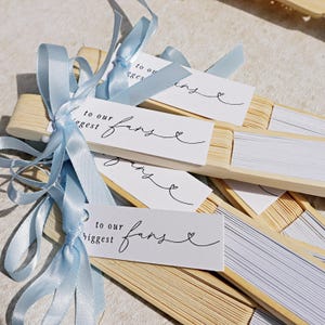 May include: A collection of paper fans with wooden frames, each tied with a light blue ribbon. White tags attached to the fans read "to our biggest fans" in black script, with a heart symbol. The fans are displayed on a light-toned surface.