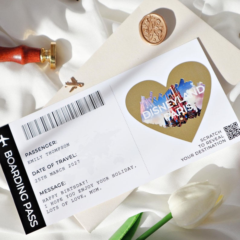 May include: A white boarding pass with a gold heart-shaped scratch-off sticker that says "Disneyland Paris". The boarding pass has the text "Passenger: Emily Thompson", "Date of Travel: 25th March 2027", and "Message: Happy Birthday! I hope you enjoy your holiday. Lots of love, Mum."