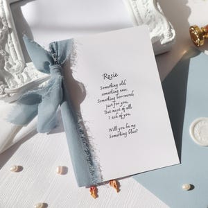May include: A white rectangular card featuring black printed text that reads: Rosie, Something old, something new, Something borrowed, just for you, But most of all, I ask of you, Will you be my Something blue? A dusty blue frayed ribbon is tied to the side.