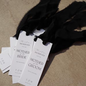 May include: White rectangular cards with black ribbon, each card has the text "This seat is reserved for the Mother of the Bride" or "This seat is reserved for the Mother of the Groom" with the names of the mothers printed below.