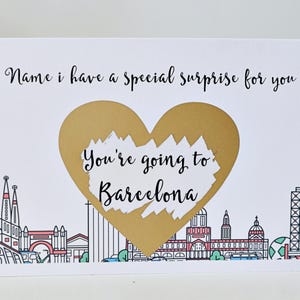 May include: A white card with a gold heart shape that says "You're going to Barcelona". The card has a black outline of the Barcelona skyline.
