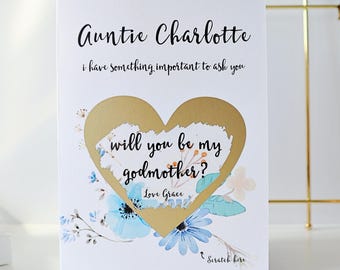 May include: A white card with gold lettering that says "Auntie Charlotte, I have something important to ask you."  A gold heart with a scratch-off area reveals the text "Will you be my godmother? Love Grace".  The card is decorated with blue and white watercolor flowers.